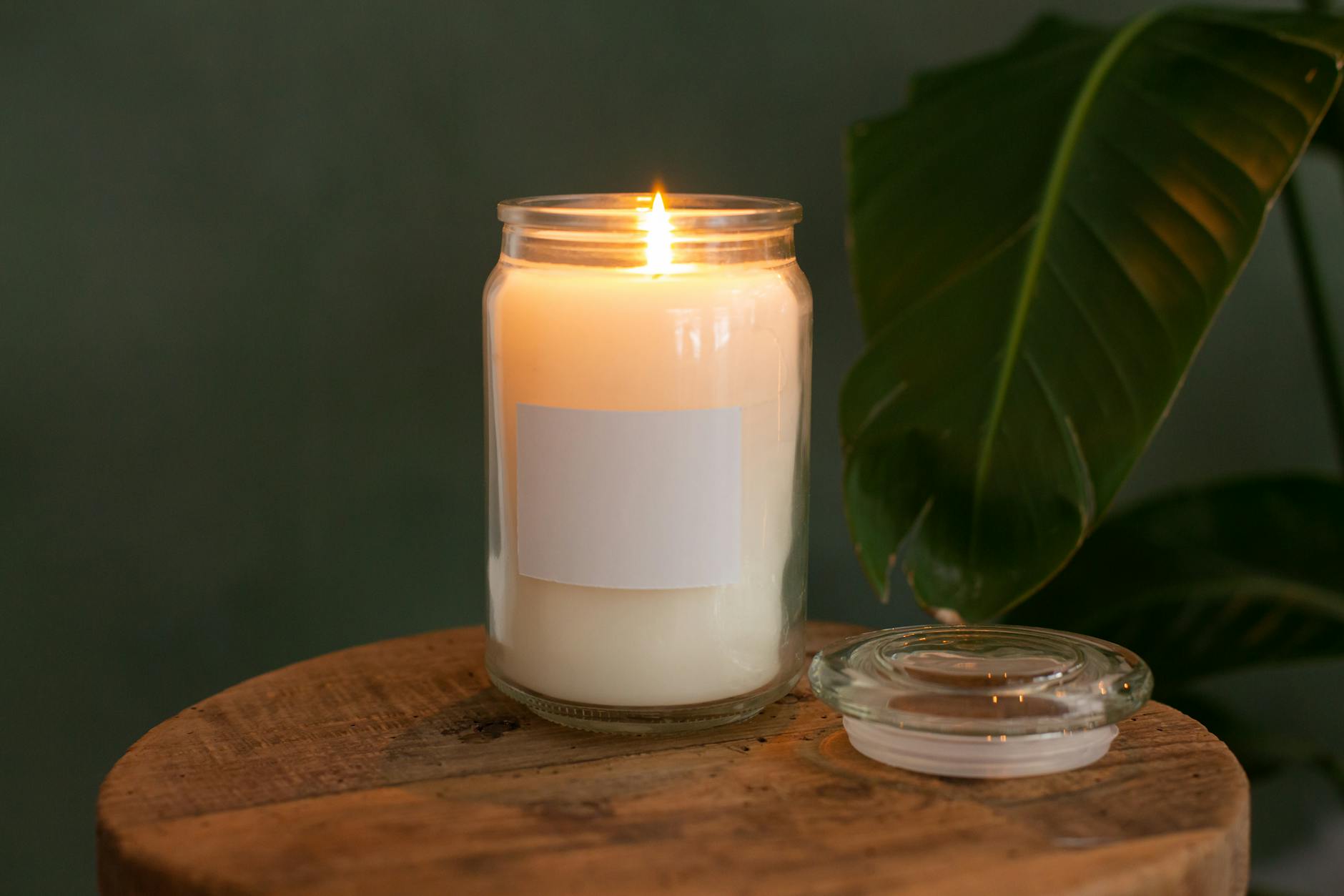 A white candle in a glass jar with a burning flame beside a green leaf on a wooden surface.
