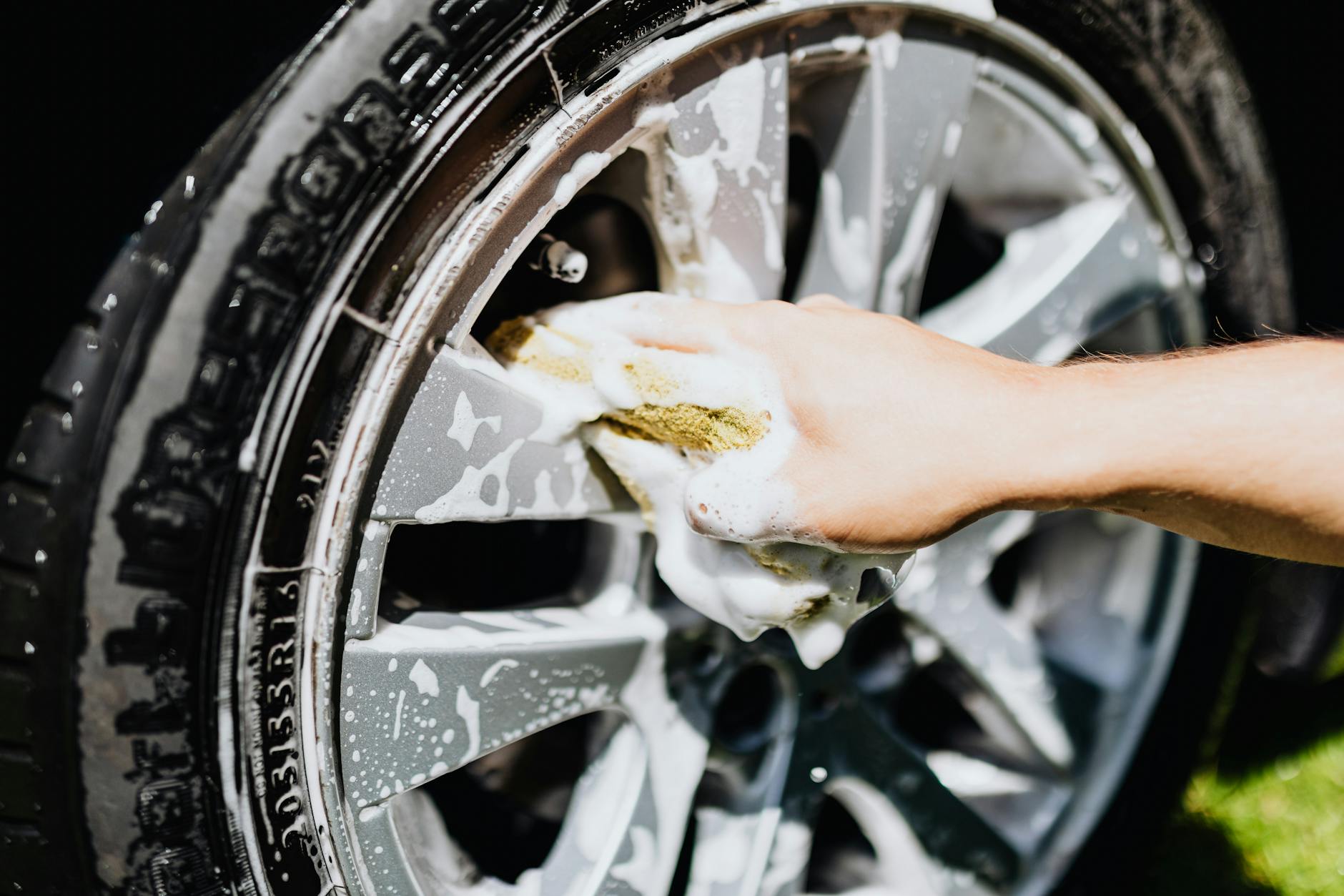 A close-up image of a hand scrubbing a car rim and tire with suds, emphasizing car wash detail.